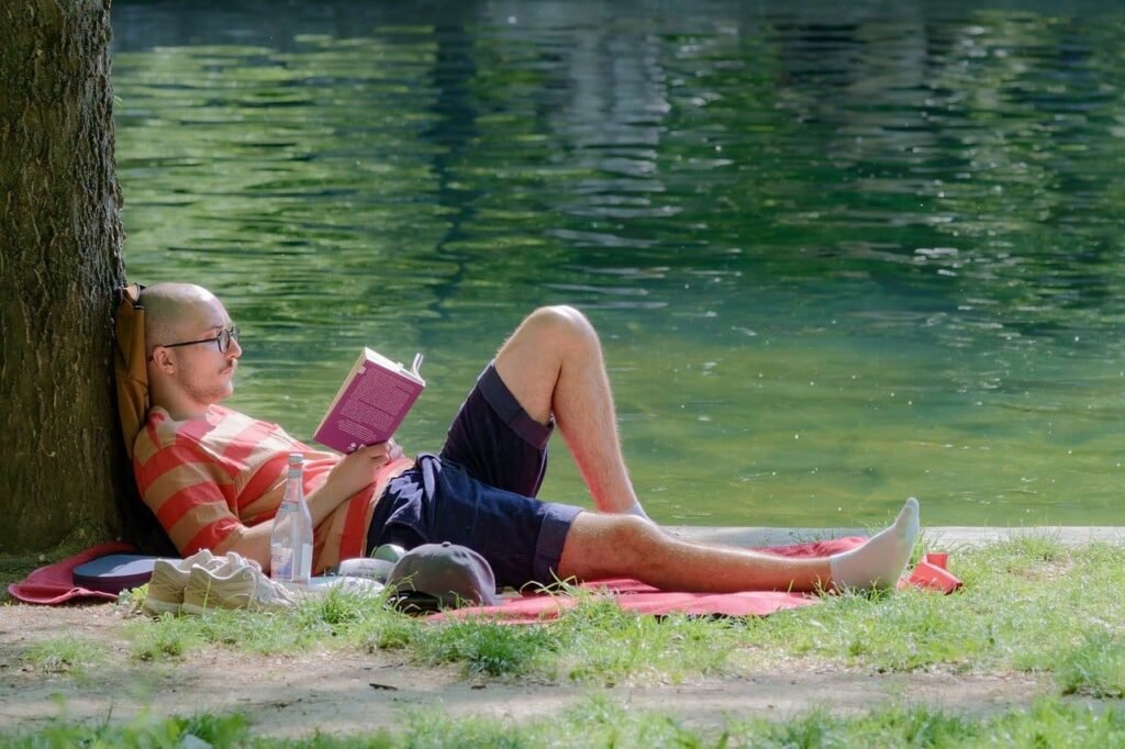 essoa deitada sobre um cobertor vermelho à beira de um lago, encostada em uma árvore e lendo um livro cor-de-rosa, com tênis, garrafa de água e boné ao lado.