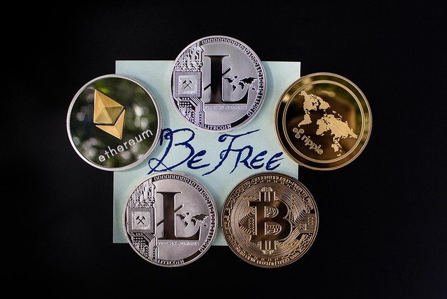 Um arranjo de cinco moedas de criptomoeda populares (Litecoin prateado, Ethereum prateado e dourado, Bitcoin dourado, e Ripple dourado) dispostas em torno de um pequeno papel quadrado de cor clara. A frase manuscrita "Be Free" (Seja Livre) está escrita em azul no centro do papel. O fundo da imagem é totalmente preto, destacando o dinheiro digital e o seu tema de liberdade financeira.