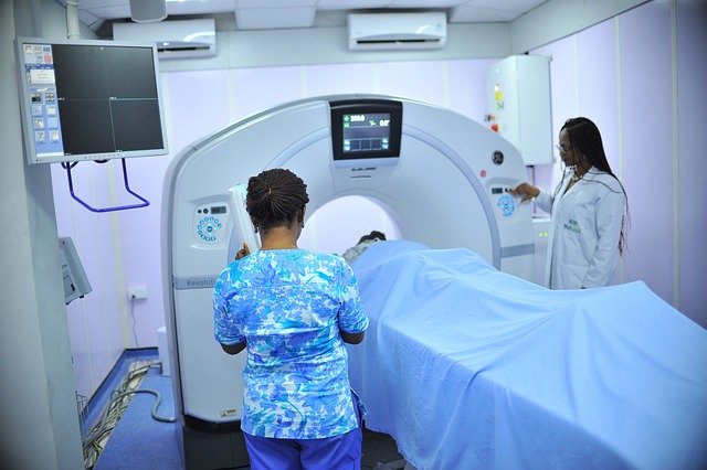 Interior de uma sala de exames com duas profissionais de saúde a operar um equipamento de tomografia computadorizada (CT). Um paciente está na mesa de exames. A imagem representa o setor de saúde ou hospitalar, que é um nicho comum de FIIs Exóticos (FIIs Hospitalares).