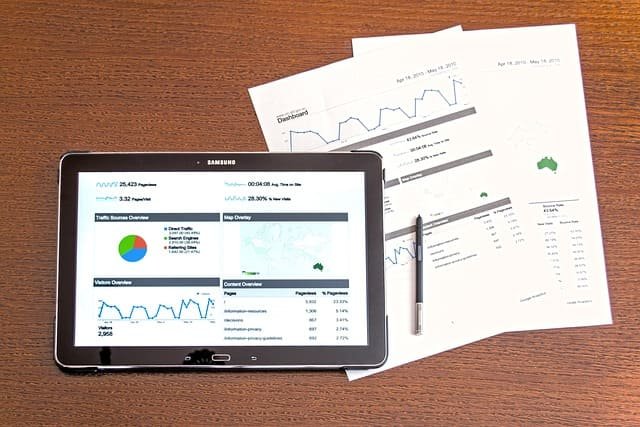 Mesa de trabalho com um tablet a exibir gráficos e dados financeiros (dashboard). O tablet e relatórios em papel, juntamente com um estilete, representam a análise e monitorização de Investimentos e desempenho de mercado.